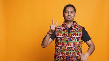 Portrait of upbeat man crossing fingers to make wish, hoping for good luck, isolated over studio background. Jolly indian person doing peace victory hand sign gesture, camera B video