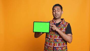 Portrait of indian man doing influencer marketing using green screen tablet, isolated over studio background. Smiling person holding empty copy space mockup device, camera B video