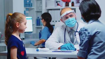 Mother explaining to doctor girl symptoms during coronavirus in medical office. Pediatrician specialist in medicine with mask providing health care services consultation, treatment in hospital cabinet video