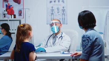 médico falando com menina dentro médico escritório e anotando a sintomas em computador portátil. especialista dentro remédio com proteção mascarar fornecendo saúde Cuidado Serviços, consulta, tratamento dentro hospital clínica. video