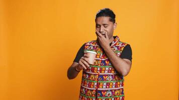 cansado indio hombre bebiendo Fresco café desde desechable papel taza temprano en el Mañana a despertar arriba y ser energizado persona Bebiendo caliente bebida desde reciclado para llevar taza, estudio fondo, cámara si video
