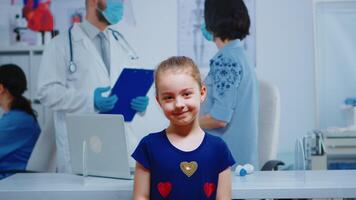 Portrait of girl smiling in medical office while mother talking with doctor in background. Specialist in medicine with protection mask providing health care services, consultation in hospital clinic video