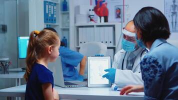 Medical specialist presenting skeleton using tablet sitting on desk in medical office. Pediatrician doctor with protection mask providing health care services, consultations, treatment during covid-19 video