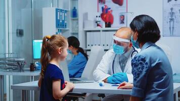 dokter controle kind temperatuur vervelend bescherming masker gedurende covid19. kinderarts specialist in geneeskunde met vizier het verstrekken van Gezondheid zorg Diensten, overleg, behandeling in ziekenhuis kastje. video