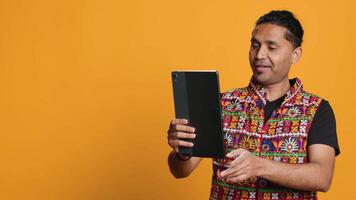 Relaxed man having entertaining conversation with friend over online videocall on device. Carefree indian person discussing with mate on teleconference using tablet, studio backdrop, camera A video