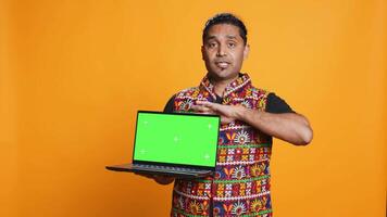 Upbeat tech specialist doing promotion using isolated screen laptop, studio background. Cheerful indian person holding chroma key notebook, doing advertisement for it, camera B video