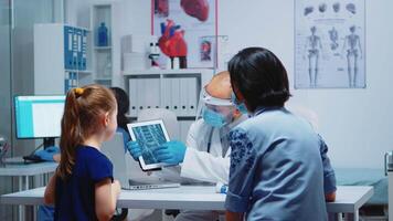 radiolog förklara röntgen använder sig av läsplatta i medicinsk kontor och sjuksköterska arbetssätt på dator. barnläkare specialist med skydd mask tillhandahålla hälsa vård service röntgen behandling undersökning video