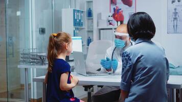 Pediatrician explaining the treatment to little girl wearing protection mask. Specialist in medicine with protection mask providing health services, consultation, treatment in hospital during covid-19 video