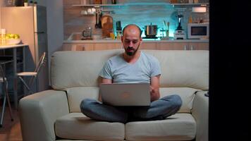 Tired man in pajams browsing on internet lifestyle information using laptop computer sitting on sofa. Freelancer working at online project in front on television late at night in kitchen video