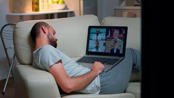 confortable homme dans pyjamas chute endormi tandis que bavardage avec collègues pendant en ligne affaires appel vidéo en utilisant portable ordinateur. caucasien Masculin séance sur canapé en retard à nuit dans cuisine video