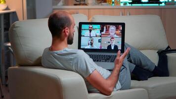 Tired man sitting comfortable on sofa while chatting with teammates about social communication during business videocall conference using laptop computer late at night in kitchen video