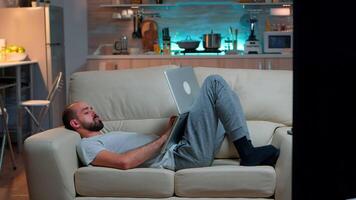 erschöpft Mann fallen schlafend während Arbeiten auf Internet Kommunikation Projekt mit Laptop Computer mit modern Technologie kabellos. kaukasisch männlich im Pyjama Sitzung auf Sofa spät beim Nacht im Küche video