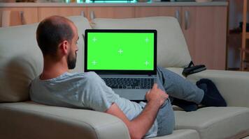 Tired man lying on sofa working on computer with mock up green screen chroma key display searching online information. Caucasian male using isolated pc late at night in kitchen video