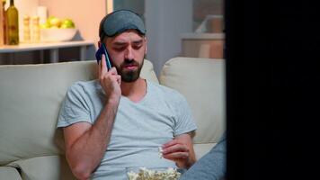 müde Mann mit Schlaf Maske stellen reden auf Smartphone Über Sozial Medien Technologie während Sitzung auf Couch und Essen Popcorn. kaukasisch männlich Aufpassen Sport zeigt an auf Fernsehen spät beim Nacht im Küche video