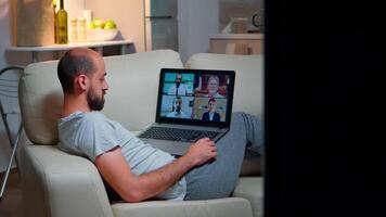 Tired man in pajamas sitting on sofa falling asleep while having online business videocall with teammates using laptop computer. Caucasian male on conference internet web communication video