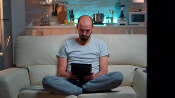 Tired man sitting alone on couch while browsing on internet using tablet computer with modern technology wireless. Caucasian male in pajamas relaxing in front of televion late at night in kitchen video