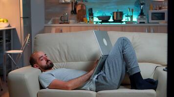 Man falling asleep in fron of TV while working on the laptop. Cozy man lying down on sofa while typing on laptop computer for social media project. Exhausted man working video