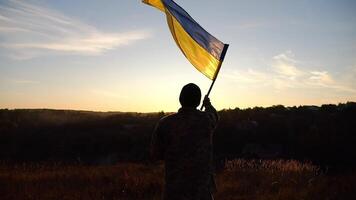 joven hombre en militar uniforme ondulación bandera de Ucrania en contra puesta de sol a antecedentes. masculino ucranio Ejército soldado levantado nacional bandera a campo. victoria en contra ruso agresión. final de guerra video