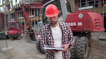 Kaukasisch Mens ingenieur architect Holding map met papieren Aan achtergrond graafmachine, bouw machinerie Bij bouw plaats. bouwer op zoek Bij bedrijf plan, bouwkundig documenten. video