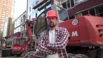 ritratto di orgoglioso costruttore a costruzione luogo. costruttore con scavatrice preparato per inizio edificio nuovo Casa. bello costruzione lavoratore in piedi nel un all'aperto la zona con pesante macchinari. ingegnere video