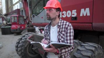 jovem construção Gerente Verificações a técnico documentação enquanto em construção local perto pesado construção equipamento trator ou escavadora. capataz verificação relatório. video