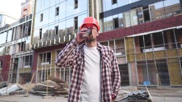 edificio supervisor Bebiendo café a sitio. arquitecto a café descanso. construcción industria trabajador en pie con café taza. hombre en protector engranaje naranja casco bebidas café a Vamos y relajarse. video