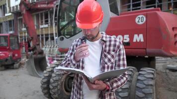 Caucasian man engineer architect holding folder with papers on background excavator, construction machinery at construction site. Builder looking at business plan, architectural documents. video