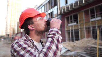 Portrait male worker in orange hard hat during rest at construction site. Man engineer hold takeaway coffee cup outside construction site in orange hard hat. Superintendent coffee break after hard day video