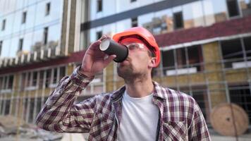 jovem caucasiano masculino capataz construtor cansado e bebidas café em fundo construção local. engenheiro dentro protetora equipamento café pausa. construção inspetor bebidas quente beber depois de Difícil dia. video