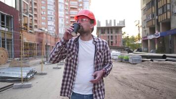 bürgerlich Ingenieur geht durch ein Baustelle und genießt Kaffee. Konstruktion Manager im schwer Hut, Plaid Shirt, und Jeans Spaziergänge Baustelle und hat Tasse von Kaffee. Architekt ruhen. Trinken heiß Kaffee zum Mitnehmen. video