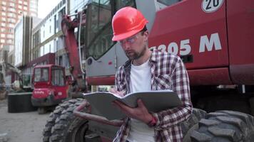 Caucasian man engineer architect holding folder with papers on background excavator, construction machinery at construction site. Builder looking at business plan, architectural documents. video