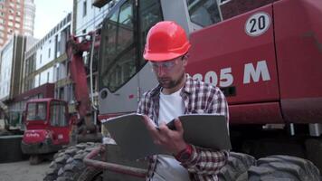 giovane costruzione manager controlli il tecnico documentazione mentre su costruzione luogo vicino pesante costruzione attrezzatura trattore o escavatore. caposquadra controllo rapporto. video