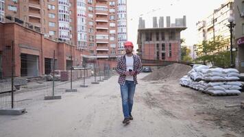 Construction engineer walking through construction site and drinking coffee. Builder in hard hat, checkered shirt and jeans walks along a building sites and has cup of coffee. Architect having break. video