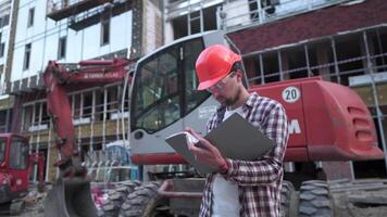 joven construcción gerente cheques el técnico documentación mientras en construcción sitio cerca pesado construcción equipo tractor o excavador. capataz comprobación informe. video