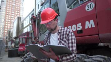 jong bouw manager cheques de technisch documentatie terwijl Aan bouw plaats in de buurt zwaar bouw uitrusting trekker of graafmachine. voorman controle rapport. video