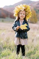 pequeño sonriente niña con un amarillo hoja en su mano y un guirnalda de otoño hojas soportes en un campo foto