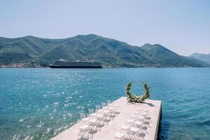 Rows of white chairs stand in front of a round wedding arch by the sea with a passing cruise ship in the background photo