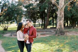 Smiling parents hugging little girl while standing with her in their arms in the park photo