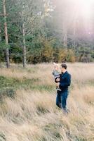 papá con un pequeño niña en su brazos camina mediante el alto césped a el borde de el bosque foto