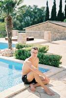 Young man sits on the shore of a swimming pool with his elbow on his knee photo