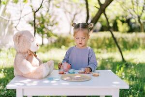 pequeño niña soportes a un mesa en el jardín y obras de teatro té fiesta con un osito de peluche oso foto