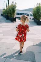 Little girl walks with a pink toy through a square with green trees in flowerpots and looks to the side. Back view photo