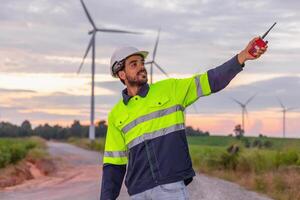 man in a yellow and black safety vest is pointing at a wind turbine. The sky is cloudy and the sun is setting photo