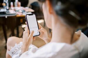 Bride in a lace robe sits on a chair and types on a smartphone. Back view photo