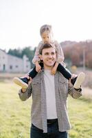 Smiling dad carries little girl on his shoulders across the meadow, looking to the side photo