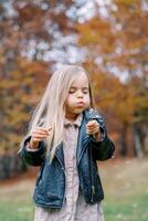 Little girl blowing on a dandelion in her hand while standing in the autumn forest photo