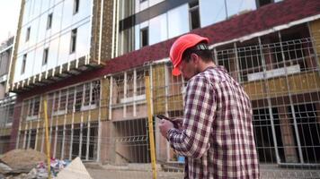 Male construction engineer walking through construction site and inspecting project, holding orange hard hat and use smart phone. Builder checking building plan on smartphone. Foreman outside at work video