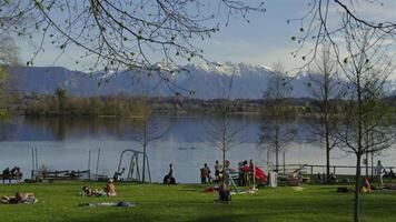 April 20, 2024. Uffing am Staffelsee, Bayern, Germany. People relaxing on green lawn by Staffelsee lake. Recreation and bathing area by am Alpenblick Uffing am Staffelsee in Bavarian Alps in spring. video