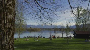 April 20, 2024. Uffing am Staffelsee, Bayern, Germany. People relaxing on green lawn by Staffelsee lake. Recreation and bathing area by am Alpenblick Uffing am Staffelsee in Bavarian Alps in spring. video