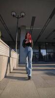 glimlachen vrouw met slank figuur wandelingen Aan stad park en teksten gebruik makend van smartphone video